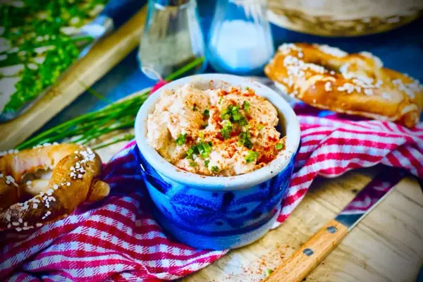 Herzhaftes Obazda-Rezept in einem blauen Steingut-Töpfchen, garniert mit Schnittlauch und Paprikapulver, daneben Brezeln und ein Messer.
